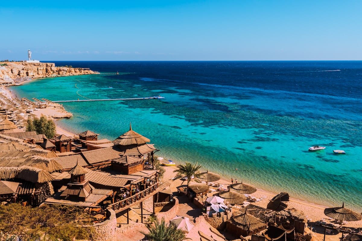 Aerial-Blick auf eine Bucht bei Sharm el-Sheikh am Roten Meer — strohgedeckte Strandrestaurants auf Stelzen, türkis-blaues Lagunenwasser, Bootsanleger und vorgelagerte Riffe.